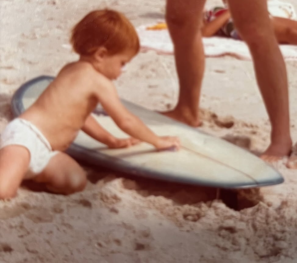 Dannon as a baby on the sand, hands on a surfboard